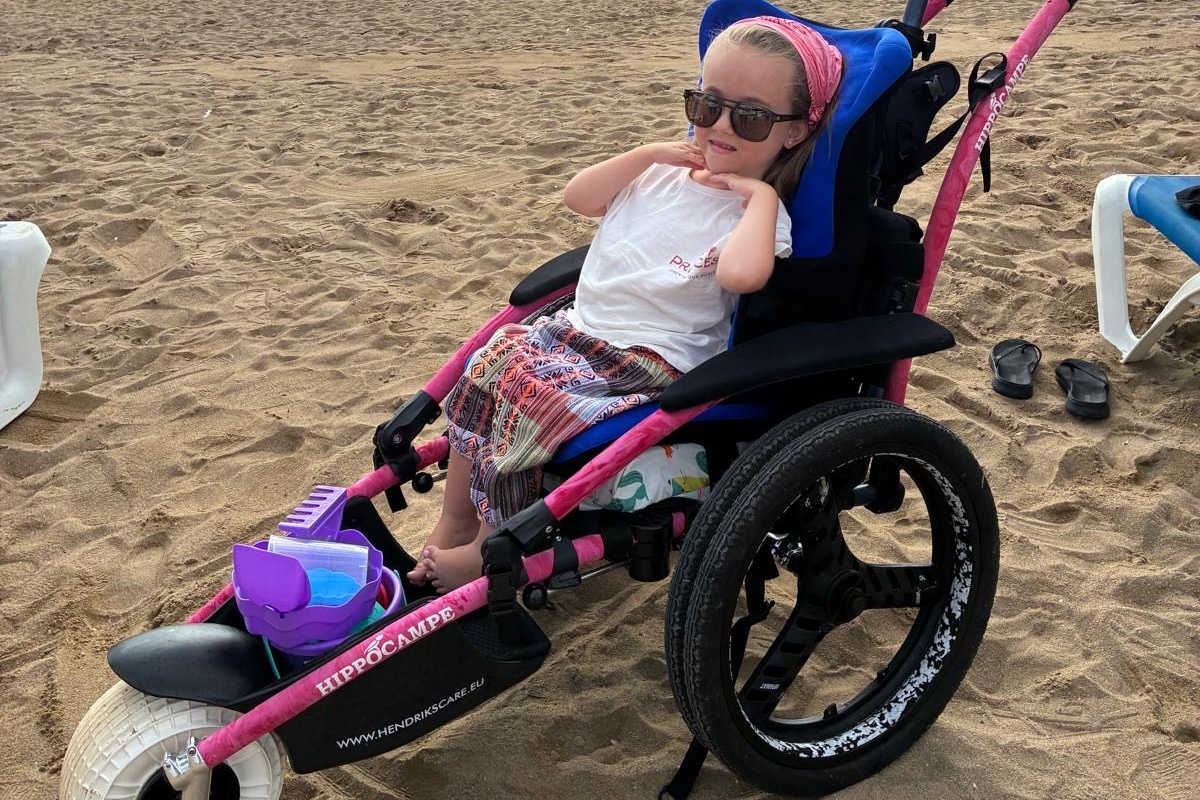 Lanah, kind-ambassadeur van Spieren voor Spieren, zit in een speciale strandrolstoel op het strand en geniet van buiten zijn ondanks haar spierziekte. title=